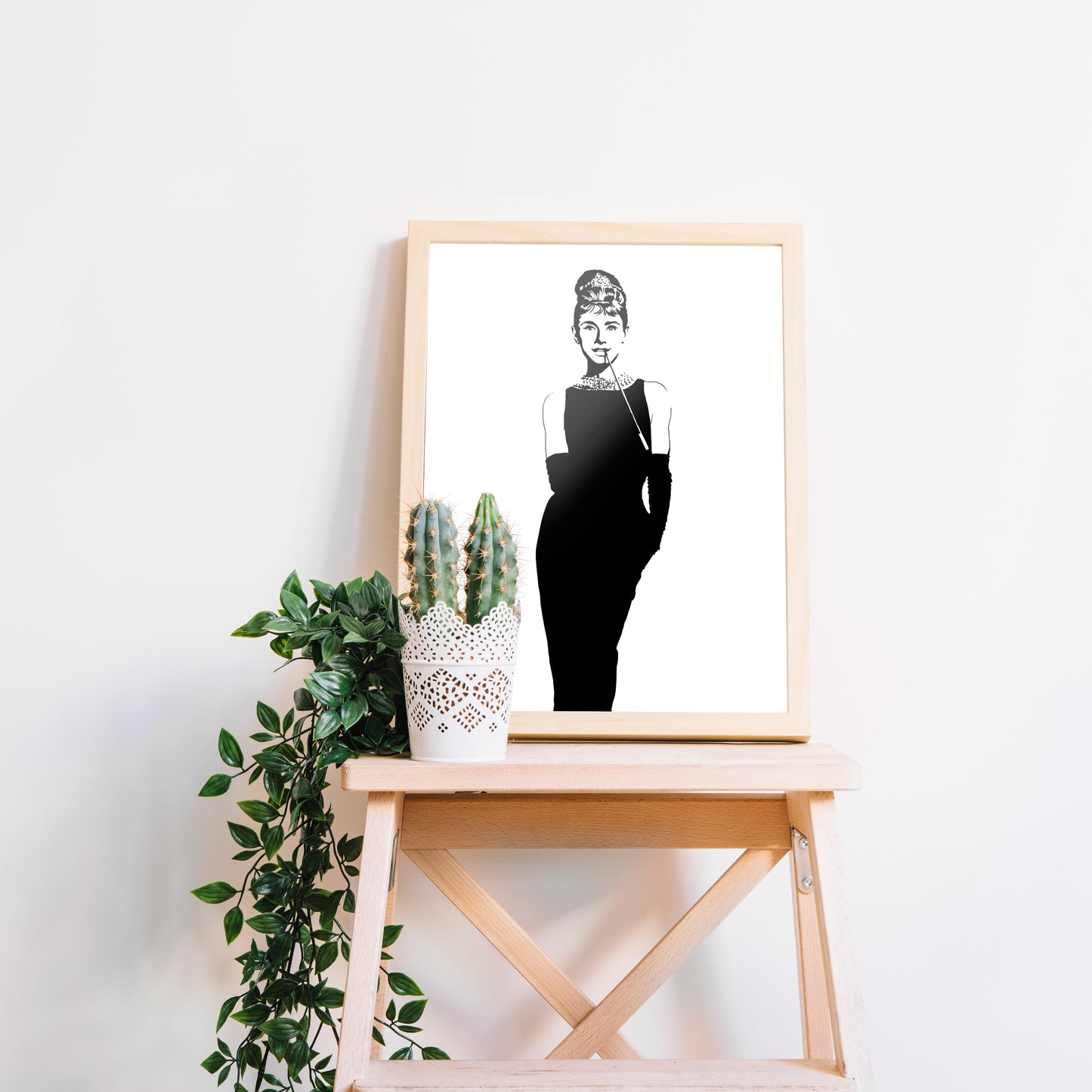 digital minimalist stencil black and white illustration of audrey hepburn as holly golightly from the film breakfast of tiffanys, slender profile in a sleek black dress and gloves to her elboys, pearl necklaces and hair in a bun with a long cigarette holder in her smiling mouth, framed print in a wooden frame on a stand next to a catcus and fern