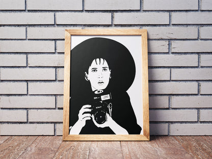 black and white illustration of lydia deetz from beetlejuice film played by winona ryder holding a nikon camera in a goth fashion outfit with a very large brimmed black hat framed art print leaned against a wall